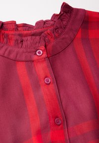 Camicia a quadri rossi con un colletto arricciato e cinque bottoni coordinati sul davanti. Il tessuto ha una consistenza liscia con un sottile luccichio.