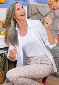 Weißes Hemd über einem weißen Tanktop, kombiniert mit hellbeigen, gemusterten Hosen. Langes graues Haar. Ananas und Früchte im Hintergrund.