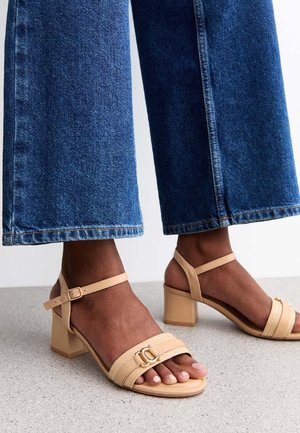 Sandali con tacco blocco beige e cinturino alla caviglia, dotati di una amplia banda per le dita e dettagli in hardware dorato, abbinati a pantaloni in denim blu a tre quarti.