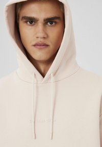 Hoodie rose clair avec cordons de serrage et un logo texturé "Serious Club" sur la poitrine. Tissu doux, design décontracté, capuche visible.