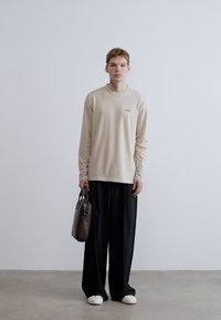 Bézs színű hosszú ujjú ing finom márkajelzéssel, laza fekete nadrággal párosítva. Egyik kézben barna táska, a fehér cipő teszi teljessé az öltözéket.