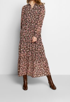 Kvinde iført en lang brun blomsterkjole med lyserøde og beige blomster, kombineret med skinnende brune ankelstøvler, stående mod en ensfarvet baggrund.