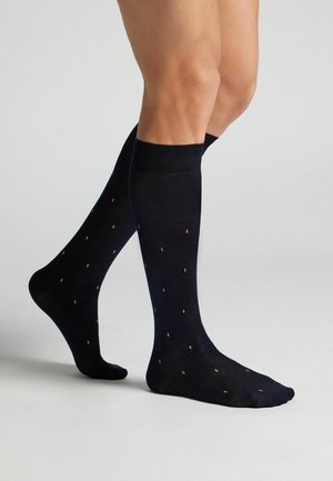 Nohy v černých nadkolenky s malými červenými a bílými puntíky, stojící na světle šedém povrchu.