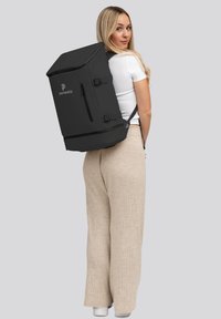 Mochila negra de forma rectangular, material de tela, bolsillo delantero con cremallera, logo en el lateral, llevada en la espalda por una persona.