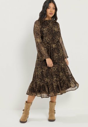 Kobieta ubrana w czarną, prześwitującą sukienkę midi z długim rękawem, z złotymi kwiatowymi wzorami oraz beżowe zamszowe botki, stojąca na jednolitym tle.