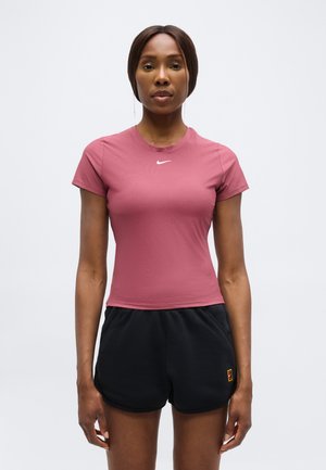 Γυναίκα με ροζ εφαρμοστό μπλουζάκι Nike και μαύρα αθλητικά σορτς, στέκεται με πρόσωπο μπροστά σε απλό φόντο.