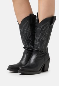 Bottes de cowboy en cuir noir avec des bouts pointus, des coutures décoratives et un talon de hauteur moyenne. Elles sont dotées de tirettes pour un enfilage facile.