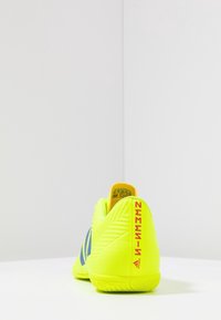 Scarpe da calcio giallo neon con strisce blu, parte posteriore testurizzata e il prominente marchio "NEMEZIZ" sul tallone. Suola in gomma con motivo a scanalatura.