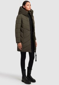 Olivgrön vadderad parkas med hög krage och chevronstickning, dragkedja, sidofickor och ett märke. Bärs med svarta leggings och stövlar.