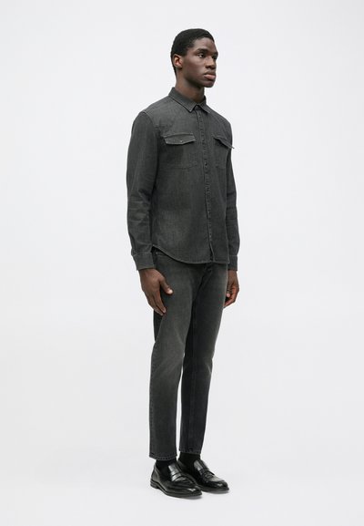 Dark denim shirt with button-down collar, two chest pockets, and seam detailing; paired with dark jeans and black loafers.