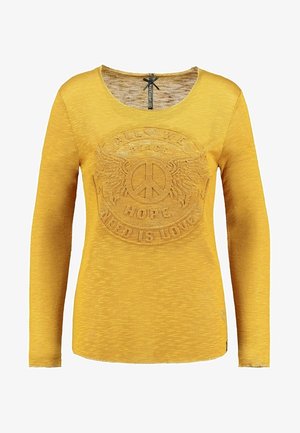 Langærmet gul skjorte lavet af struktureret stof med et forhøjet fredssymbol med vinger og tekst: "Alt vi har brug for er kærlighed."