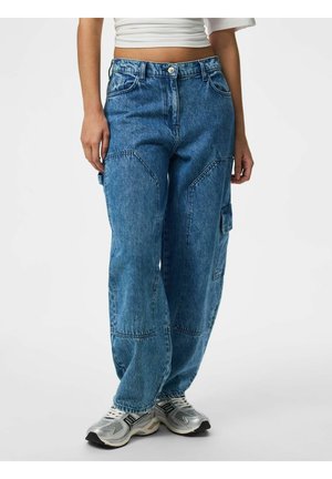 Femme portant un jean cargo ample en denim bleu avec poches plaquées et des chaussures de sport argentées sur un fond blanc uni.