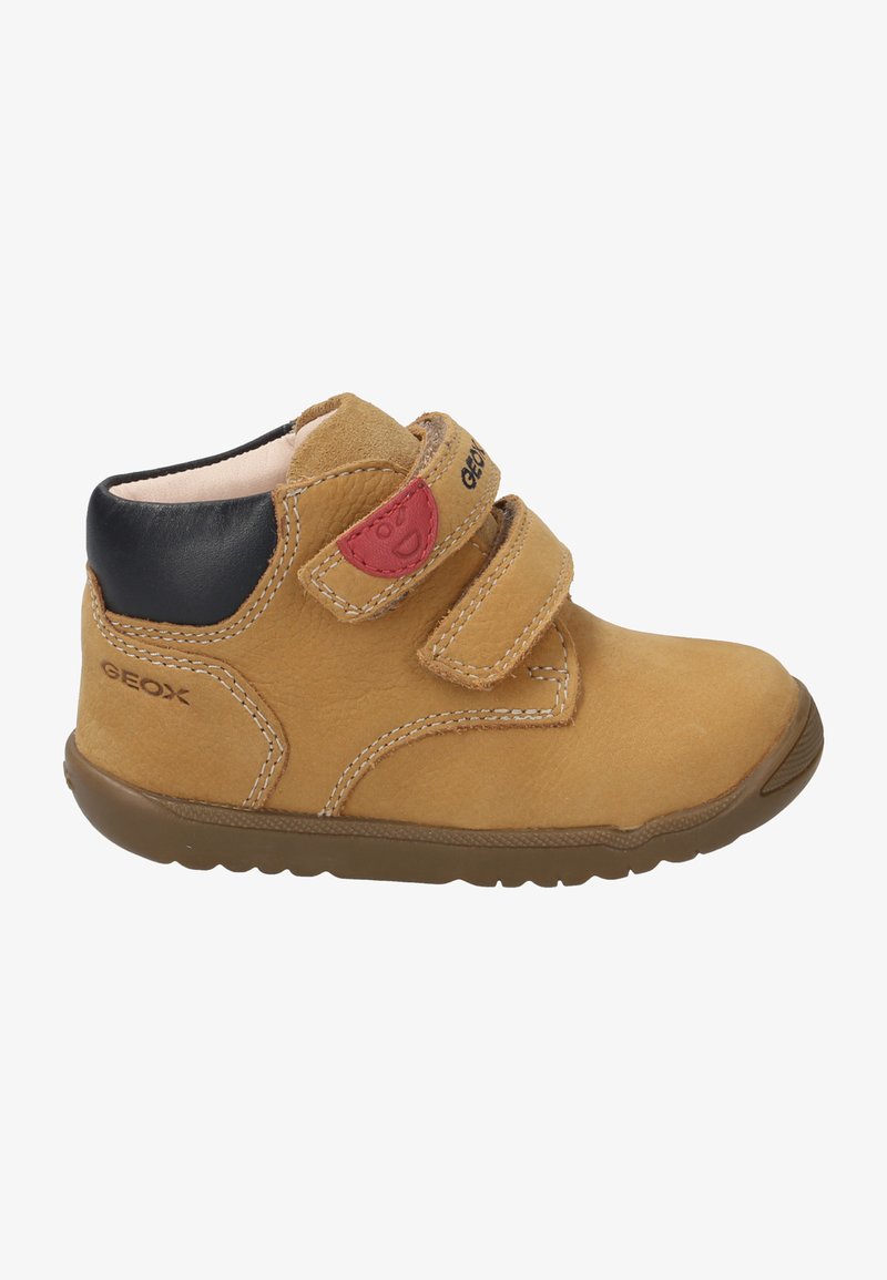 Bottine en cuir tan avec un talon noir, deux sangles ajustables en Velcro, des détails de couture courbés et une semelle en caoutchouc. Accent rouge près des sangles.