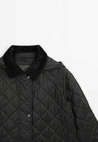 Gewatteerd zwart jack met een corduroy kraag en capuchon. Voorzien van drukknopen en een geometrisch diamantenpatroon op de stof.