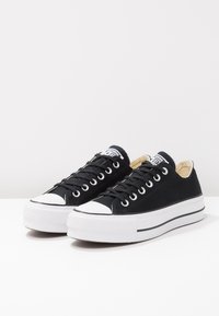Schwarze Canvas-Sneaker mit niedrigem Schnitt, weißen Gummizähnen, Schnürdesign, flachen weißen Sohlen und kontrastierenden weißen Nähten.