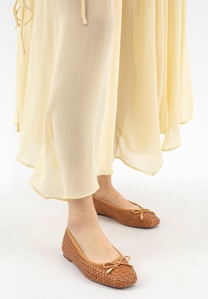 Voeten met bruine gevlochten ballerina's met strikjes, onder de zoom van een vloeiende lichtbeige rok of jurk.