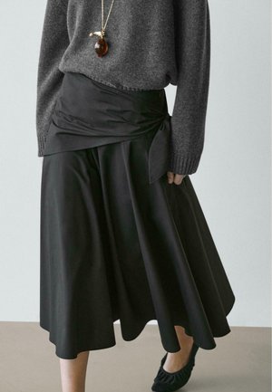 Jupe portefeuille noire à la texture lisse et satinée, associée à un pull gris et des chaussures plates noires à volants. Un collier pendentif en or vient souligner l'ensemble.