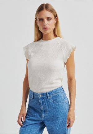 Kermanvärinen teksturoidulla pinnalla varustettu toppi, jossa lyhyet liehuvat hihat, yhdistetty vaaleansinisiin farkkuihin, joissa on hienovarainen pilkkukuosi.