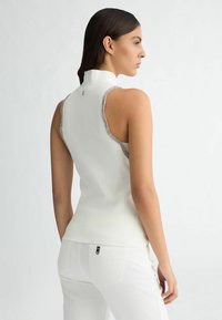 Top bianco senza maniche con colletto alto, materiale texturizzato e rifiniture argentate lungo le aperture per le braccia, abbinato a pantaloni bianchi.