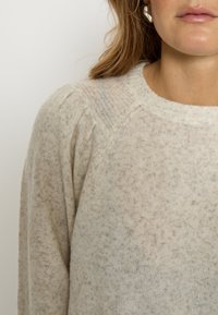 Svetlo béžový sveter z jemnej, textúrovanej látky. Má okrúhly výstrih, raglánové rukávy a celoplošný bodkovaný vzor.