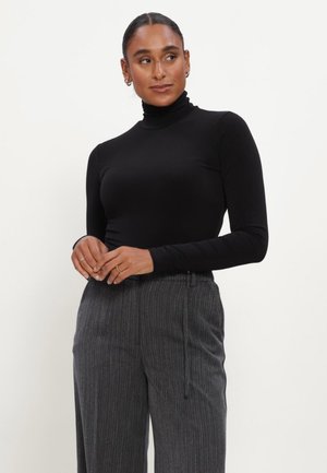 Vrouw met naar achteren gebonden haar, gekleed in een zwarte coltrui en grijze broek met krijtstrepen, staand tegen een effen lichte achtergrond.