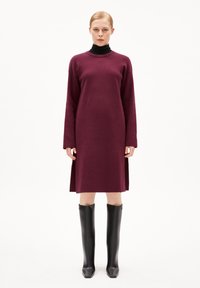 Abito lungo bordeaux a maniche lunghe con colletto alto, fessure laterali e una vestibilità ampia, abbinato a stivali neri lucidi al ginocchio.