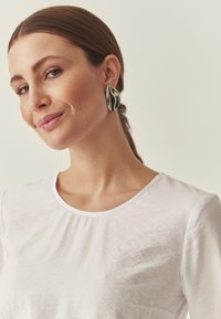 T-shirt blanc léger avec un col rond porté par une personne. Grandes boucles d'oreilles argentées brillantes avec un design abstrait.