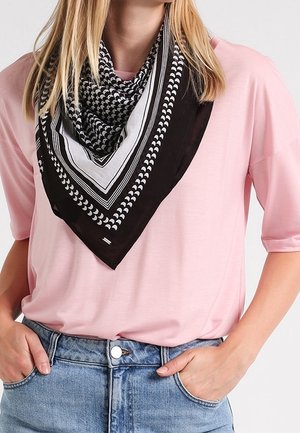 Femme blonde portant une écharpe noire et blanche à motifs sur un t-shirt rose à manches courtes, rentrée dans un jean bleu, les mains dans les poches.