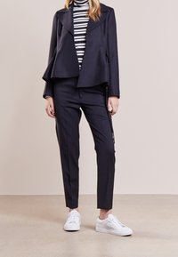 Giacca navy doppiopetto con orlo a campana, dolcevita a righe, pantaloni neri su misura e sneakers bianche. Tessuto liscio e design aderente.