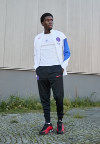 Hvid jakke med blå accenter, sorte Nike bukser og sneakers med røde accenter. Har runde logoer og en afslappet pasform. Tekstileret stof.