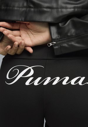 Manos cruzadas detrás de la espalda con una chaqueta de cuero negra, enfocándose en los pantalones negros con un gran logo blanco de "Puma" en la parte trasera.