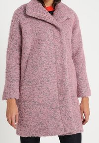 Manteau en laine rose texturée à manches longues avec grand col porté sur un pantalon noir et un haut rouge.
