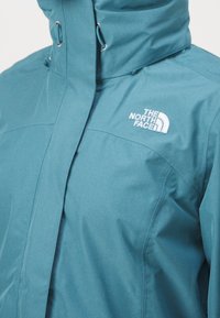 Veste imperméable bleue avec col montant et logo "The North Face" blanc brodé sur la poitrine, dotée de cordons de capuche réglables.