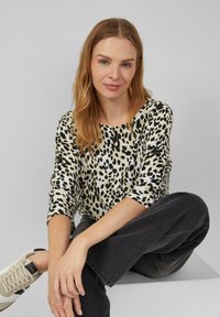 Leopardenmuster-Oberteil in Beige und Schwarz, mit Dreiviertelärmeln und Rundhalsausschnitt, kombiniert mit dunklen Jeans und hellen Sneakers.