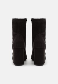 Un par de botas de tobillo de ante negro con tacones de bloque vistas desde la parte posterior, con costuras visibles y tiradores de cremallera.