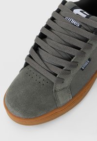 Šedé semišové skateboardové topánky s hrubými šedými šnúrkami, perforovanou oblasťou špičky, gumenou podrážkou a logom "etnies" na jazyku a bočnej nášivke.