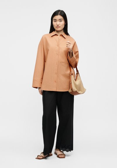 Žena nosí voľnú hnedú košeľu na gombíky, čierne široké nohavice s krajkovými lemami, hnedé sandále a drží béžovú pletenú kabelku.