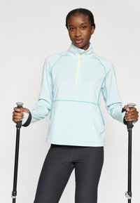 Ανοιχτό γαλάζιο αθλητικό μπλουζάκι μακρυμάνικο με ημιδιάφανο φερμουάρ στο γιακά, ραφές σε αντίθετο τιρκουάζ χρώμα και θηλιές για τους αντίχειρες, συνδυασμένο με σκούρο κολάν και ραβδιά πεζοπορίας.