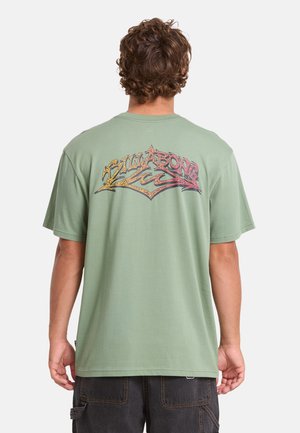 Uomo con capelli ricci che indossa una maglietta verde chiaro con una grafica colorata "Billabong" sulla schiena, abbinata a pantaloncini cargo scuri.
