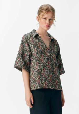 Blumenbluse in Schwarz, aus einem weichen Stoff mit kurzen Ärmeln, Kragen und einem lebhaften Muster aus rosa und grünen Blumen.