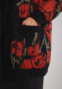 Cárdigan de punto con base negra, que presenta patrones florales rojos y acentos verdes. Incluye una textura acanalada y detalles de bolsillo en la parte frontal.