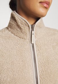 Beżowa polarowa kurtka z wysokim kołnierzem, z gładkim zamkiem z przodu i teksturowanym wykończeniem dla ciepła i komfortu.