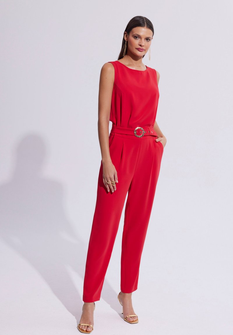 Czerwonakombinezon bez rękawów o dopasowanej sylwetce, ozdobny pasek z okrągłym akcentem oraz zwężane nogawki. Gładki materiał, brak widocznych wzorów.