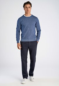 Blauer gestrickter Pullover mit Rundhals-Ausschnitt, kombiniert mit dunkelblauen Hosen und weißen Sneakers. Das Model steht vor einem hellen Hintergrund.