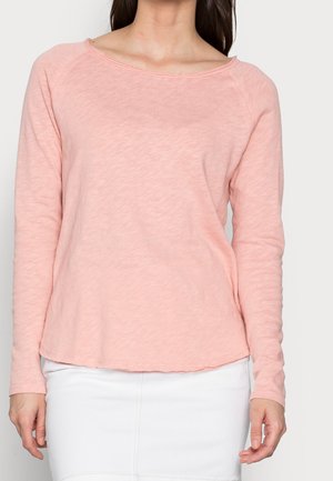 Vrouw die een lichtroze top met lange mouwen en een witte rok draagt, getoond van de nek tot halverwege de dij tegen een effen achtergrond.