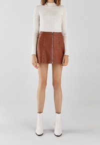 Hnědá mini sukně z umělé kůže s předním zipem, texturovaným povrchem a dvěma bočními kapsami. Kombinována s přiléhavým bílým topem s žebrováním a bílými kotníkovými botami.