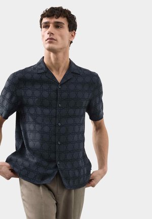 Man draagt een marineblauw overhemd met korte mouwen met een subtiel geometrisch patroon en beige broek, staat voor een effen lichte achtergrond.