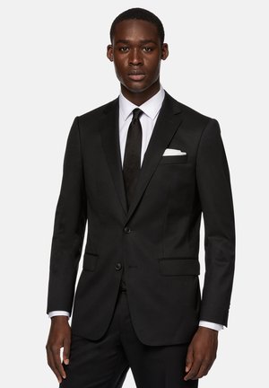 Schwarzer maßgeschneiderter Anzug mit Reverskragen, zwei Knöpfen und Taschen. Weißes Hemd und schwarze Krawatte, mit einem Einstecktuch in der Brusttasche.