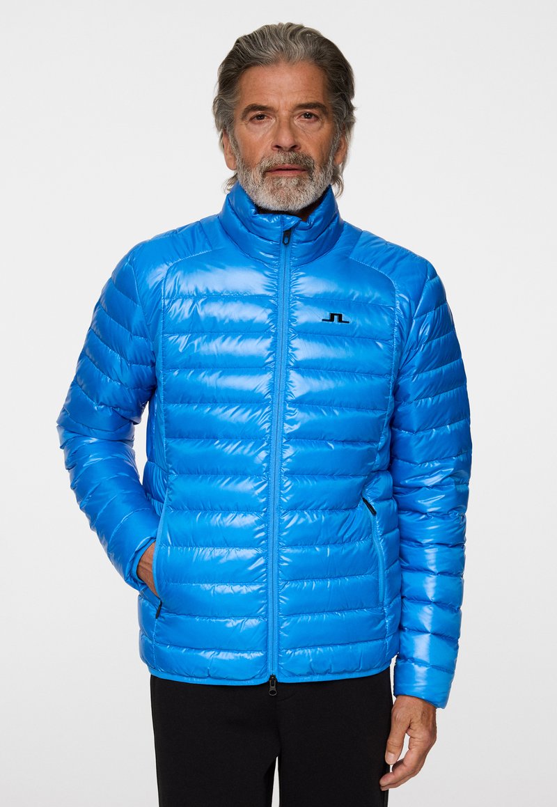 Blå puffjacka med horisontella quiltade sektioner, hög krage, dragkedja fram och sidofickor; glänsande yta med svart logotypdetalj.