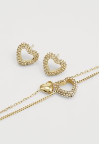 Orecchini e bracciale a forma di cuore in tonalità oro, decorati con strass trasparenti. Il bracciale presenta una chiusura a cuore con logo incisa.